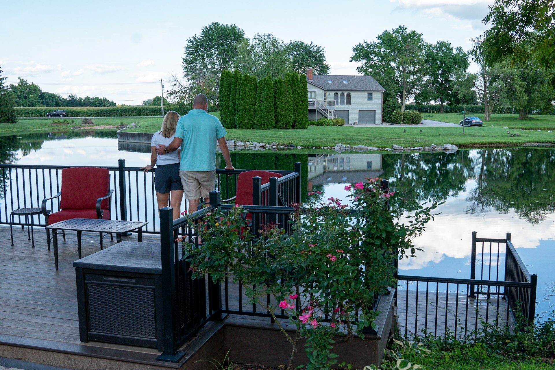 A couple stands arm-in-arm on a landscaped wooden deck overlooking a calm pond. The deck features black metal railings and outdoor furniture, including red cushioned chairs and a small table. A bush with pink flowers grows nearby, while across the pond, a modern two-story house with a lawn, trees, and a parked car is reflected in the still water under a partly cloudy sky.