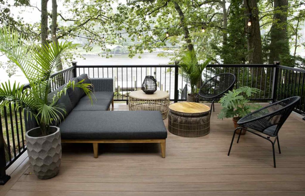 An outdoor deck surrounded by black railing featuring modern patio furniture and overlooking a wooded lakeside setting — showcasing a stylish outdoor living space by Fence and Deck Depot