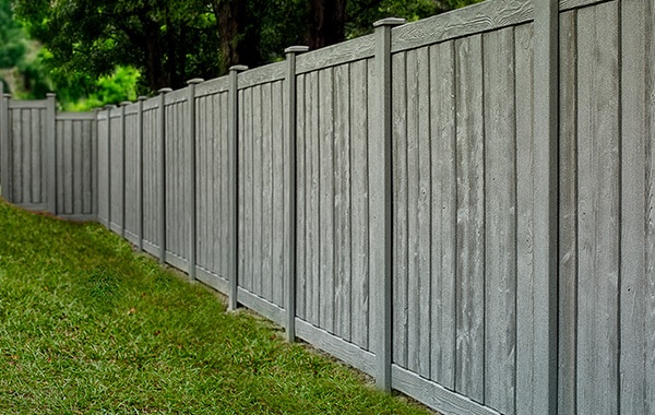A tall composite privacy fence surrounds a grassy yard. The fence is grey and looks similar to wood.