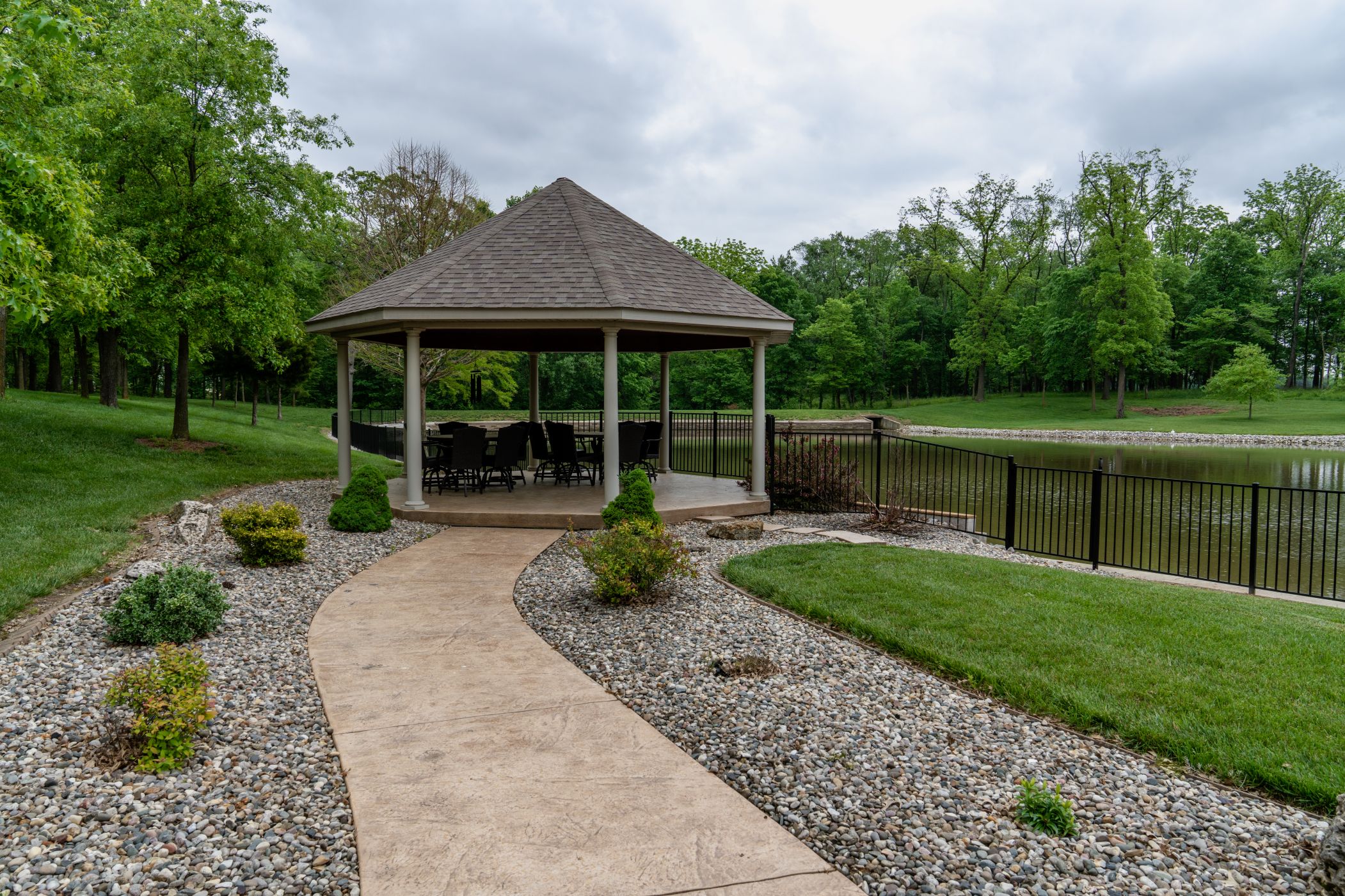 A large yard with a covered seating area is enclosed by a black aluminum fence. A large pond is on the other side of the fence.