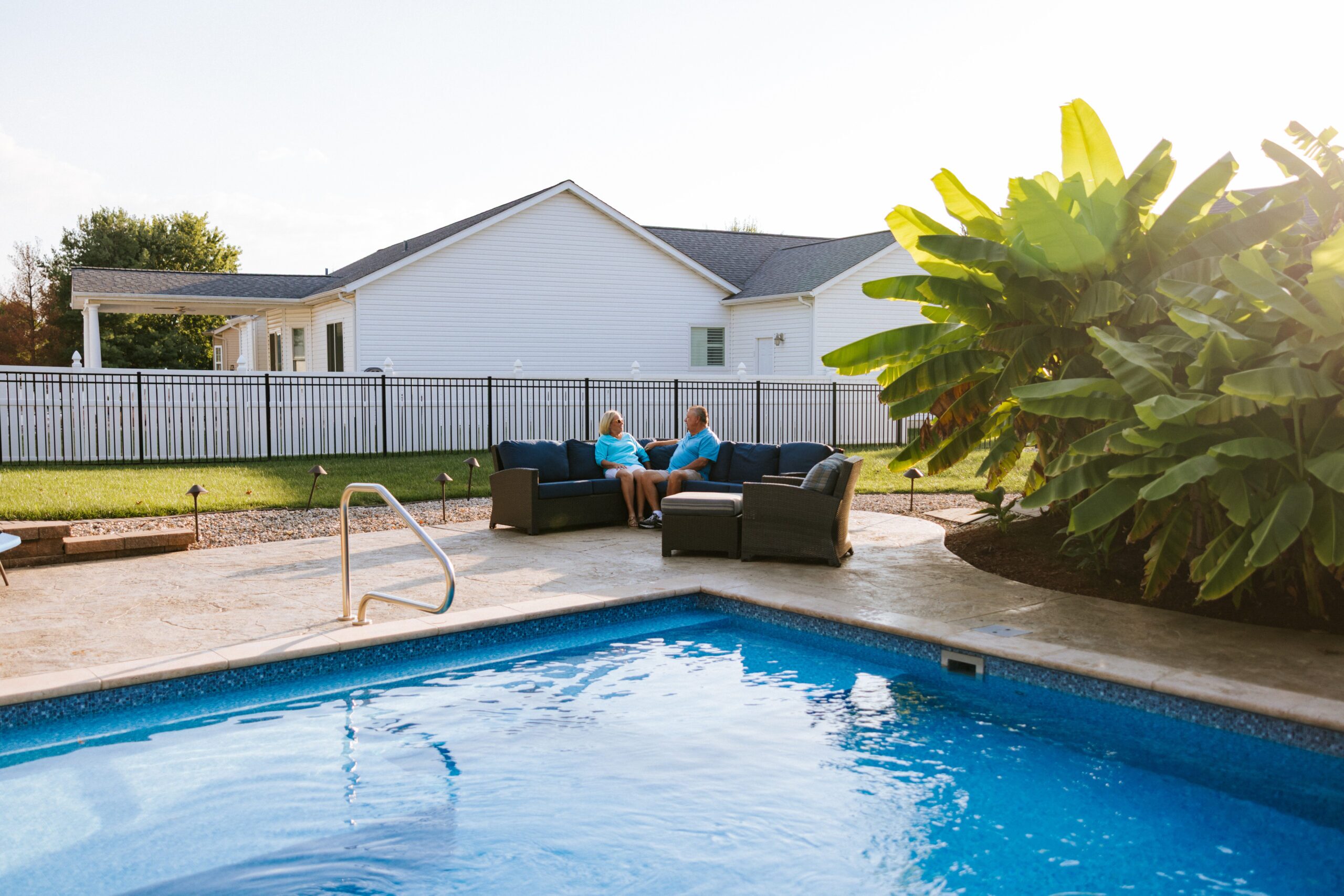 Couple sits on patio furniture inside fenced-in pool area. The fence is black aluminum and surrounds the yard.