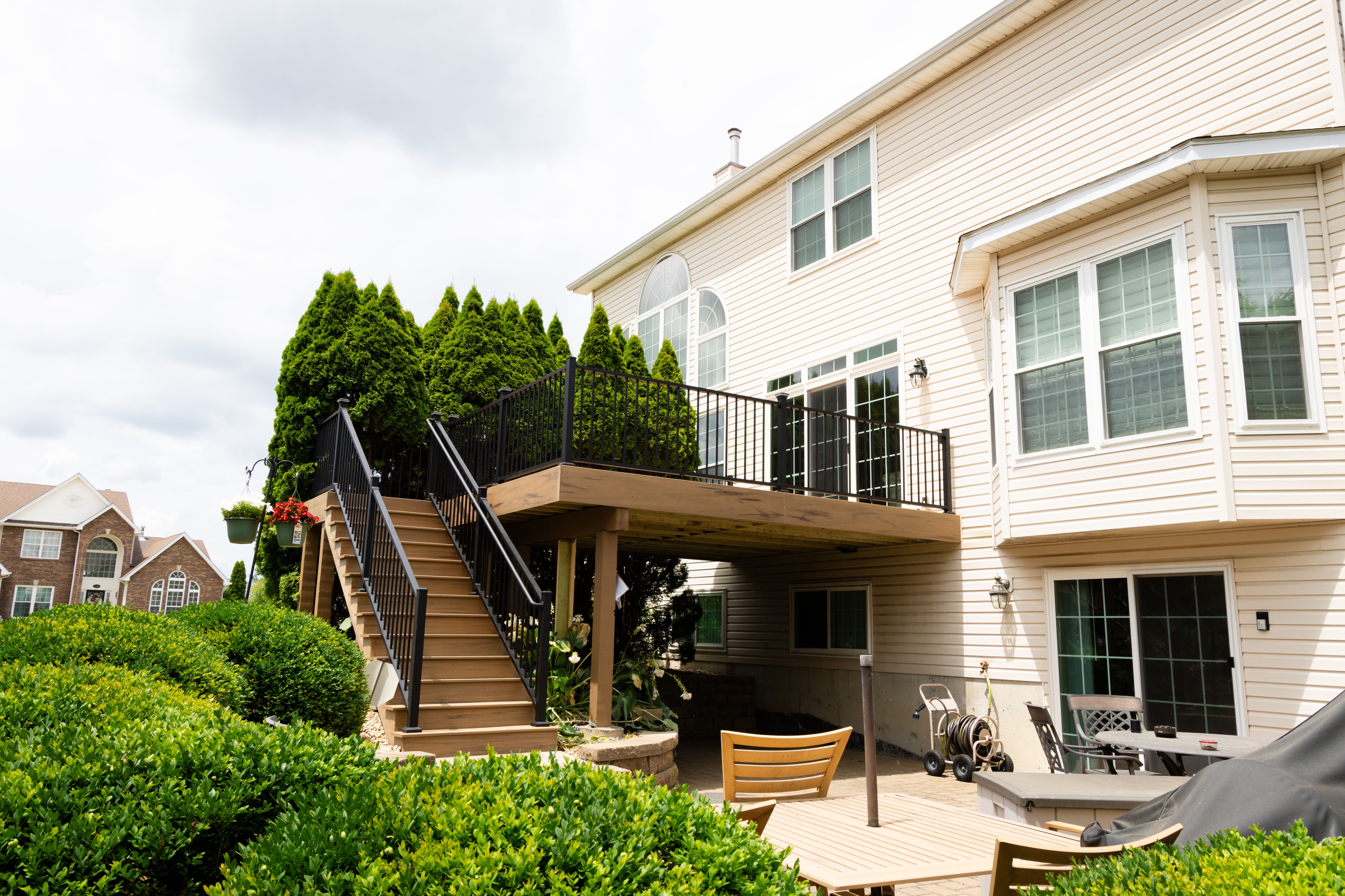 Backyard view of a two-story beige house with a raised wooden deck featuring black railings and stairs, surrounded by tall evergreens and landscaped shrubs, with outdoor furniture and patio space below