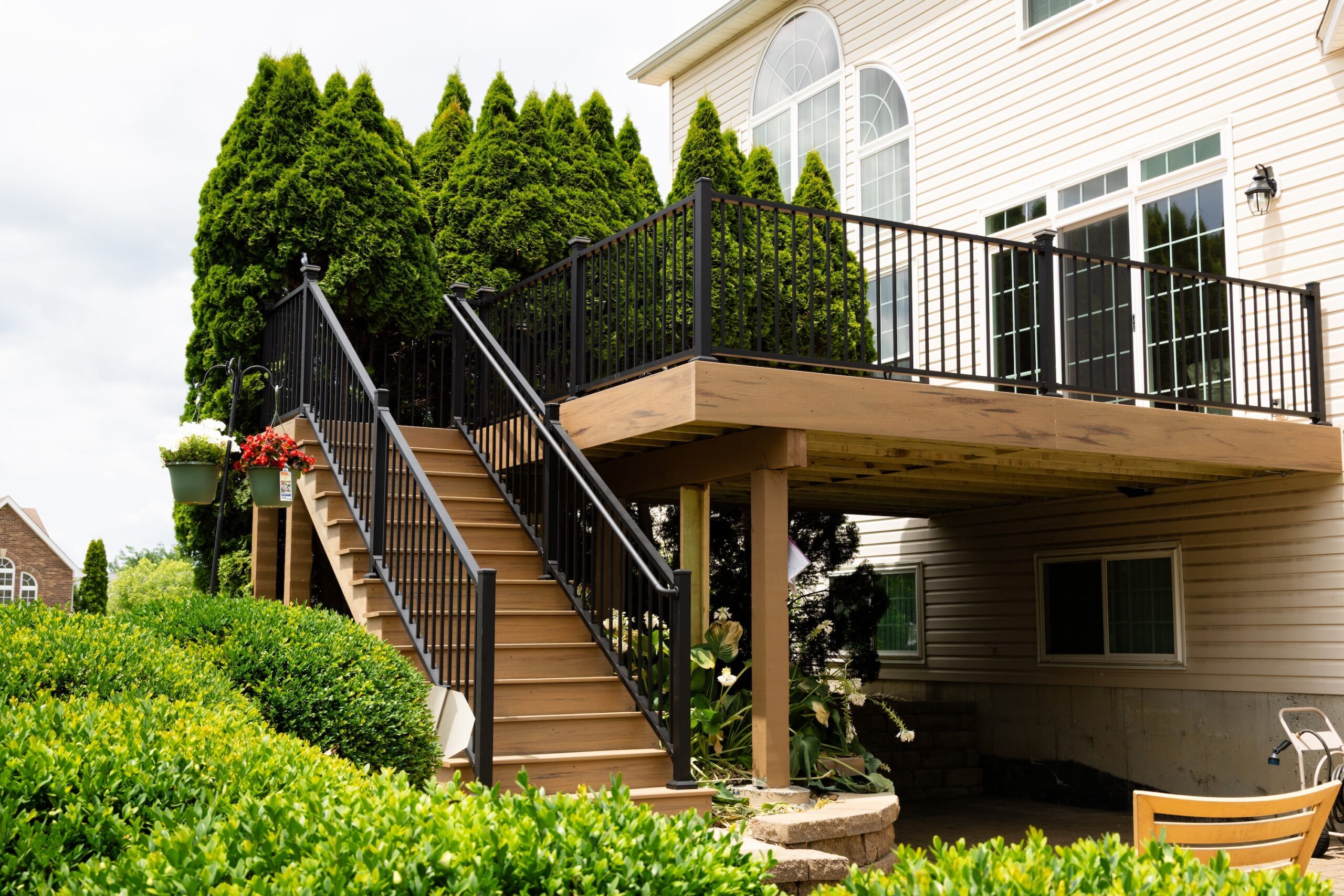 Elevated backyard deck by Fence Deck and Depot with black metal railings and wooden stairs surrounded by lush green landscaping, attached to a beige house with large windows