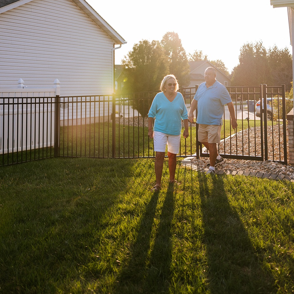 A couple walks in to their fenced-in yard. The fence is black aluminum and has a gate for entry.