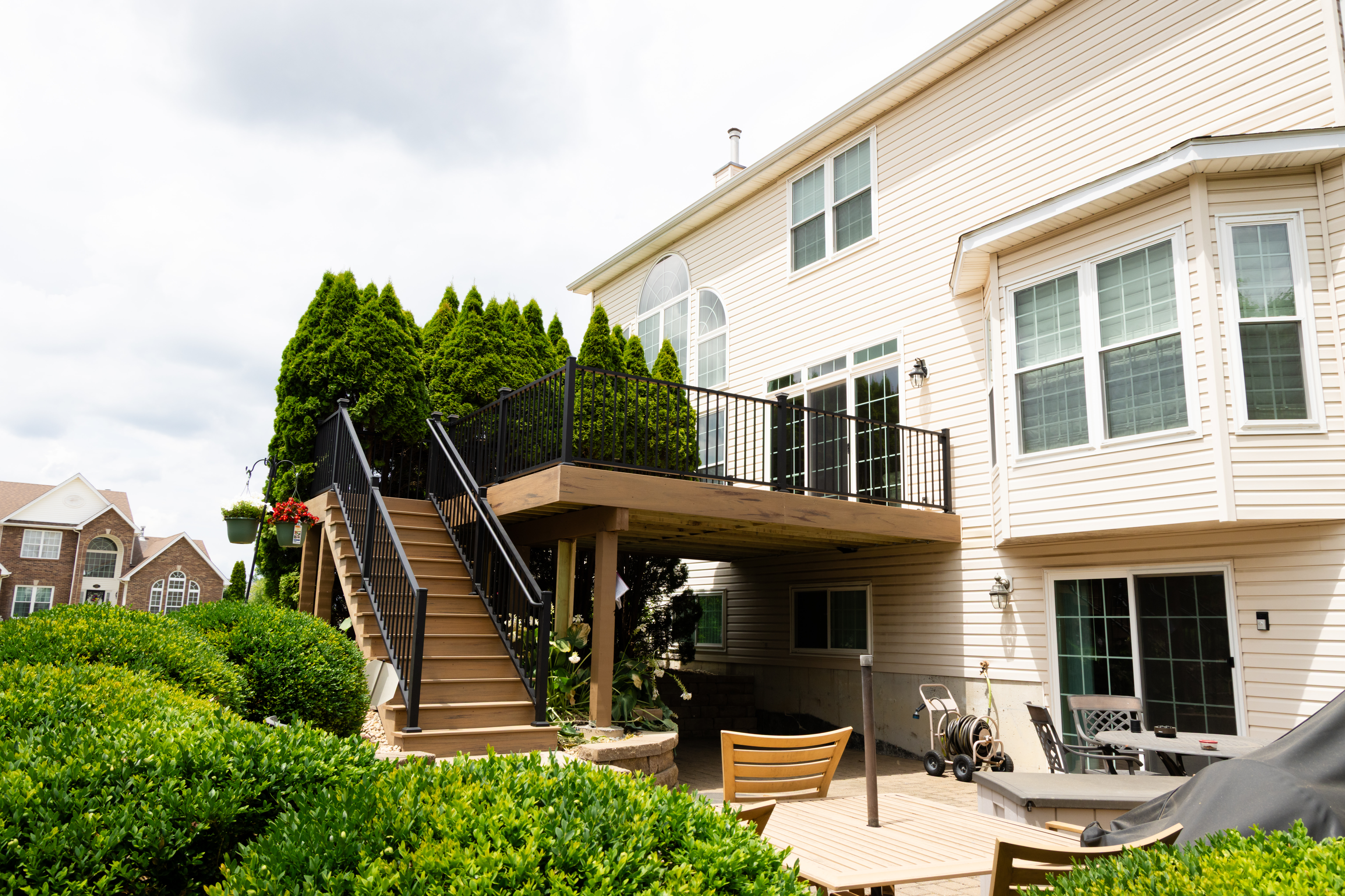 Backyard view of a two-story beige house with a raised wooden deck featuring black railings and stairs, surrounded by tall evergreens and landscaped shrubs, with outdoor furniture and patio space below
